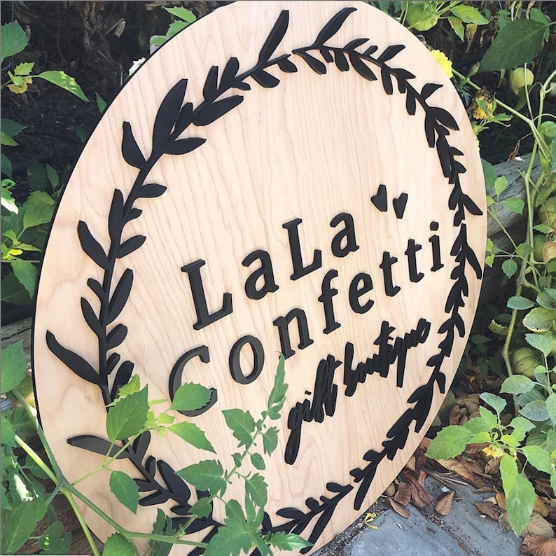 Puede incluir: Un letrero redondo de madera con un dise&ntilde;o de corona negra y el texto "LaLa Confetti gift boutique".