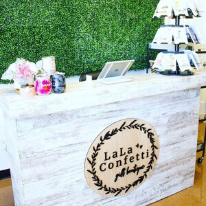 Puede incluir: Un mostrador de madera blanco con un letrero que dice "LaLa Confetti gift boutique" en letras negras. El letrero es un c&iacute;rculo de madera redondo con un dise&ntilde;o de corona alrededor del texto.