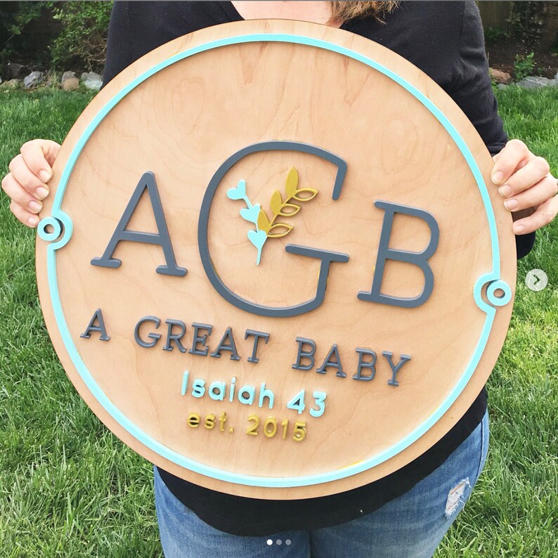 Puede incluir: Un letrero redondo de madera con un borde azul. El letrero dice "AGB A Great Baby Isaiah 43 est. 2015". Las letras est&aacute;n en una fuente negra con un contorno blanco. Hay un dise&ntilde;o de hoja de oro en el centro del letrero.