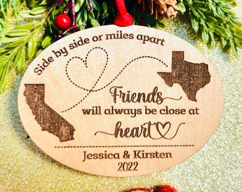 May include: Wooden ornament with a heart design and the text "Side by side or miles apart Friends will always be close at heart Jessica & Kirsten 2022". The ornament features outlines of California and Texas.