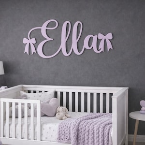May include: A lavender wooden name sign that reads "Ella" in cursive script, with bow accents. The sign is mounted on a gray wall above a white crib with lavender bedding. The room has a soft, feminine aesthetic.