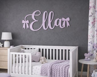 May include: A lavender wooden name sign that reads "Ella" in cursive script, with bow accents. The sign is mounted on a gray wall above a white crib with lavender bedding. The room has a soft, feminine aesthetic.