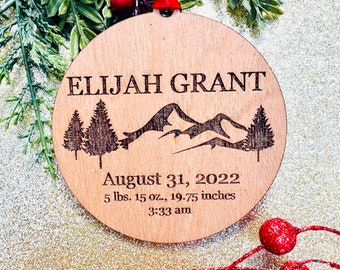 May include: A round wooden ornament with a mountain and pine tree design. It is personalized with the name "ELIJAH GRANT" and the birth date "August 31, 2022". The ornament also includes the birth weight and length: "5 lbs. 15 oz., 19.75 inches" and the time of birth: "3:33 am".