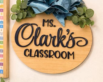 May include: A wooden classroom sign with the text "Ms. Clark's Classroom" in navy blue script. The sign is round and adorned with a teal bow and faux greenery. A framed chart is visible on the left.