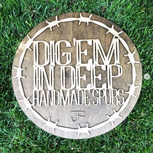 Puede incluir: Un letrero redondo de madera con un borde de alambre de p&uacute;as. El letrero dice "Dig 'Em In Deep Handmade Spurs" con un monograma "J" en el centro inferior.