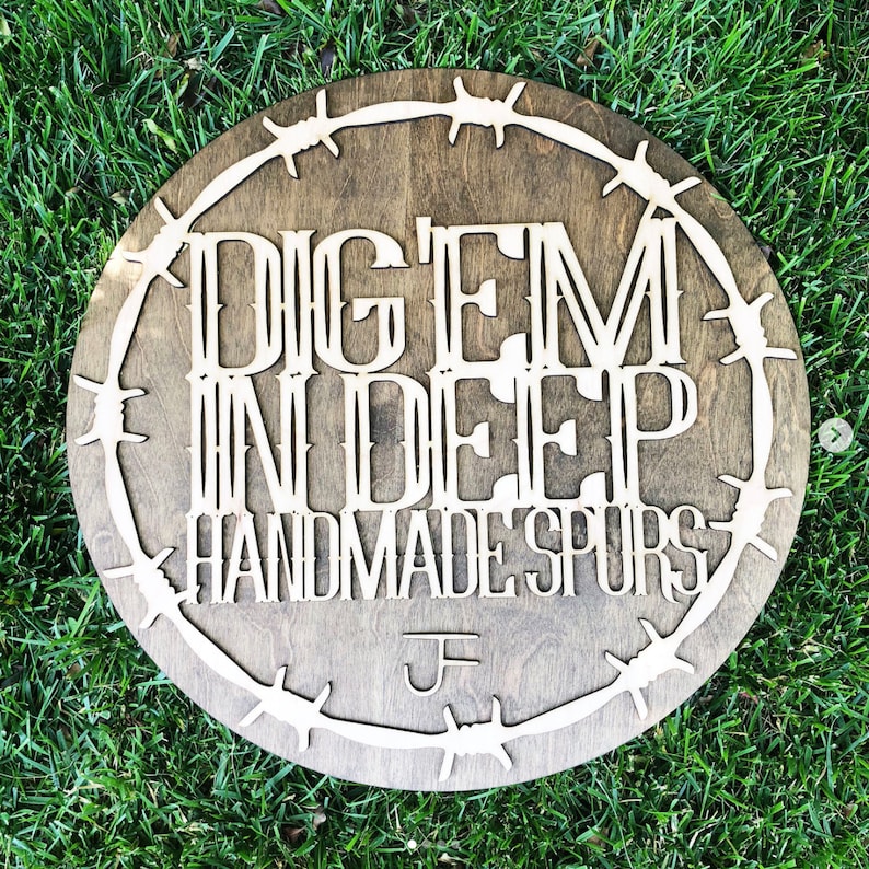 Puede incluir: Un letrero redondo de madera con un borde de alambre de p&uacute;as. El letrero dice "Dig 'Em In Deep Handmade Spurs" con un monograma "J" en el centro inferior.