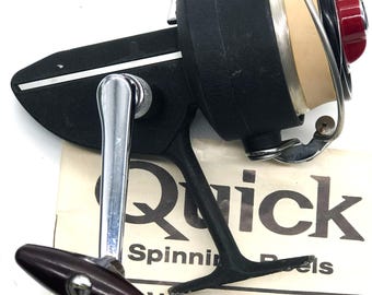 May include: Black and silver fishing reel with a red knob and a beige spool. The reel has a silver handle and is labeled "Quick Spinning Reels". A paper with "WARRANTY and REPAIR STATIONS" is beneath the reel.