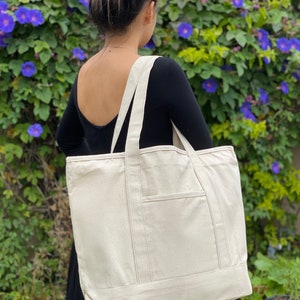 May include: A large, off-white canvas tote bag with a single pocket on the front. The bag has two long straps and is made of a sturdy, natural fabric.