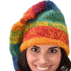 Puede incluir: Una mujer lleva un gorro y una bufanda de punto de colores. El gorro tiene una cola larga que le cuelga de la espalda. La bufanda está envuelta alrededor de su cuello y anudada en la parte delantera. Ella sonríe y mira a la cámara.