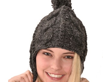 Puede incluir: Un gorro de punto gris con un pompón en la parte superior y orejeras. El gorro tiene un patrón de punto de cable y lo lleva puesto una persona con el pelo rubio.