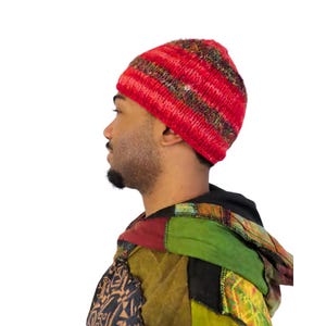 May include: A red knit beanie with horizontal stripes of varying colors. The hat is made of a soft, textured material. The person is wearing a patchwork jacket with a black hood.