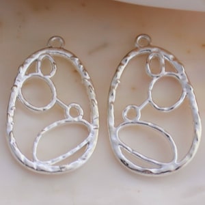 May include: Two silver-colored, openwork teardrop-shaped earring pendants. Each pendant features an abstract design of interconnected circles and ovals within a textured frame. The pendants have a small loop at the top for attachment.