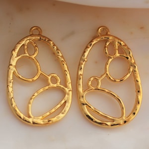 May include: Two gold-tone metal earring charms with an oval shape and a cutout design of circles and ovals.
