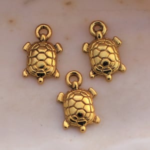 May include: Three gold-colored turtle charms with a detailed shell design. The charms are perfect for jewelry making or crafting.
