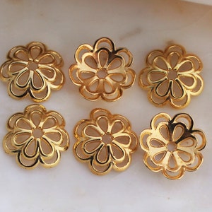 May include: Six gold-colored, flower-shaped jewelry components. Each component has a detailed, openwork design with petal-like shapes. The components are arranged on a light-colored surface, likely for crafting or jewelry making.