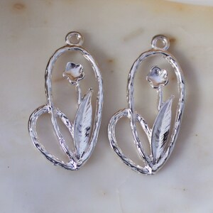 May include: Two silver-toned metal charms in the shape of a flower with a heart-shaped design. The charms have a delicate, intricate design and are perfect for adding a touch of elegance to jewelry projects.
