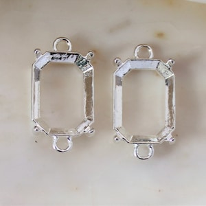 May include: Two silver-toned metal bezel settings for jewelry making. Each setting has a rectangular shape with rounded corners and two loops on the bottom for attaching to a chain or cord.
