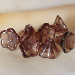 May include: Close-up of several translucent, flower-shaped beads in a deep plum color. The beads have a delicate, petal-like design with a small hole in the center, perfect for stringing. The beads are clustered together, showcasing their unique shape and color.