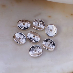 May include: A collection of silver-colored, flat, round beads with a sunburst design radiating from a central hole. The beads are scattered on a light-colored, textured surface.
