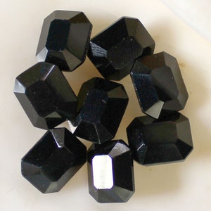 May include: Eight black faceted glass beads, each with a rectangular shape. The beads are arranged in a cluster on a white surface.