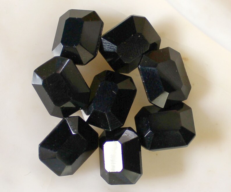 May include: Eight black faceted glass beads, each with a rectangular shape. The beads are arranged in a cluster on a white surface.