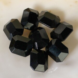 May include: A group of eight black faceted glass beads, each with a rectangular shape. The beads are arranged in a cluster on a white surface.
