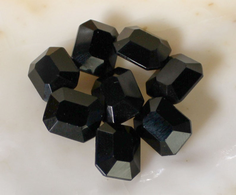 May include: A group of eight black faceted glass beads, each with a rectangular shape. The beads are arranged in a cluster on a white surface.