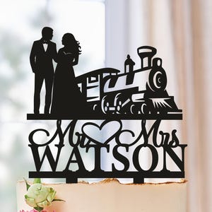 Pode incluir: Um bolo de casamento com um cake topper de silhueta preta com noivos, um comboio e as palavras "Mr & Mrs Watson". O bolo é decorado com flores rosa e brancas e está sobre um prato branco.