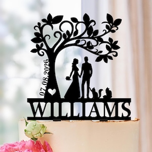 May include: Black laser-cut wedding cake topper featuring a tree design with a heart, a couple holding hands, and two cats. The name "WILLIAMS" is displayed below the couple, with the date "07.08.2026" on the tree trunk.