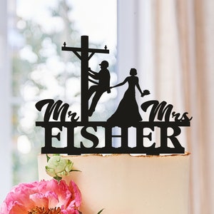 May include: A wedding cake with a black silhouette cake topper featuring a bride and groom, with the groom on a utility pole. The topper reads "Mr. & Mrs. FISHER". The cake is decorated with pink and white flowers.
