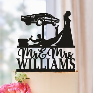 May include: Black silhouette cake topper featuring a bride and groom. The groom is lying on his back with a car on a lift above him. The bride is standing with her foot on the groom's leg. The text "Mr & Mrs WILLIAMS" is below the figures.