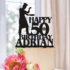 May include: Black silhouette cake topper of a chef holding a pan with the text "Happy 50th Birthday Adrian" on a white cake with pink flowers.