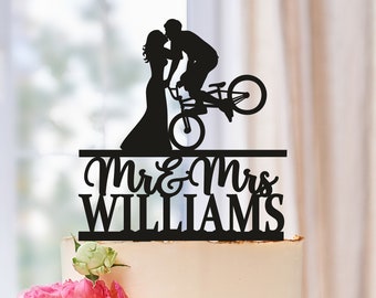 May include: Black silhouette of a couple kissing on a bicycle, with the text "Mr & Mrs WILLIAMS" below. The topper is on a white cake with pink flowers.
