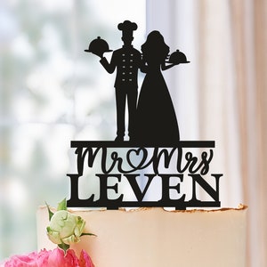 Può includere: Topper per torta in silhouette nera di una sposa e uno sposo. Lo sposo indossa un cappello da chef e tiene un vassoio da portata. La sposa tiene un vassoio da portata. Il topper dice "Mr & Mrs LEVEN" in un carattere decorativo.
