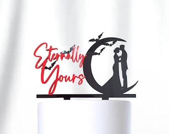 Può includere: Un cake topper nero e rosso con le parole "Eternally Yours" e una silhouette di una coppia che si bacia sotto una luna crescente con pipistrelli. Il topper è posto su una torta bianca a più strati con un nastro nero.