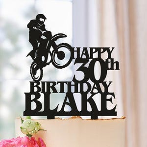 May include: A birthday cake with a black cake topper featuring a silhouette of a motocross rider and the words "HAPPY 30th BIRTHDAY BLAKE." The cake is decorated with pink and white flowers and sits on a white plate.