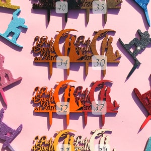 May include: Ten wooden cake toppers with the text "I Put a Spell on You" and a couple standing under a crescent moon. The toppers are in various colors, including black, brown, orange, red, and natural wood.