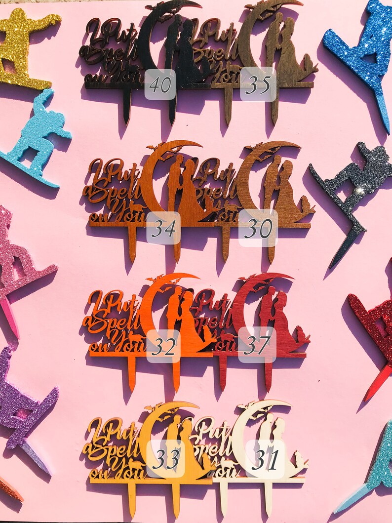 May include: Ten wooden cake toppers with the text "I Put a Spell on You" and a couple standing under a crescent moon. The toppers are in various colors, including black, brown, orange, red, and natural wood.
