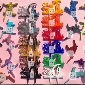 May include: A set of 39 acrylic cake toppers in various colors and designs. The toppers feature a couple under a crescent moon, a snowboarder, and the text "But first, we spell it out".