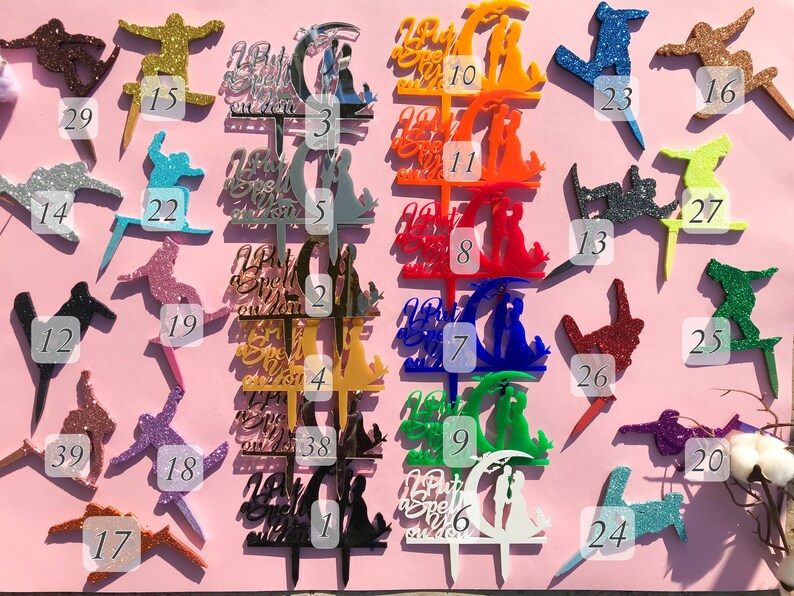 May include: A set of 39 acrylic cake toppers in various colors and designs. The toppers feature a couple under a crescent moon, a snowboarder, and the text "But first, we spell it out".