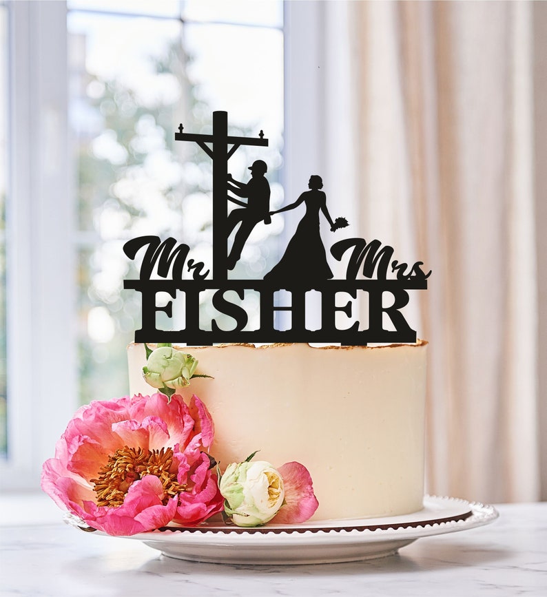 May include: A wedding cake with a black silhouette cake topper featuring a bride and groom, with the groom on a utility pole. The topper reads "Mr. & Mrs. FISHER". The cake is decorated with pink and white flowers.
