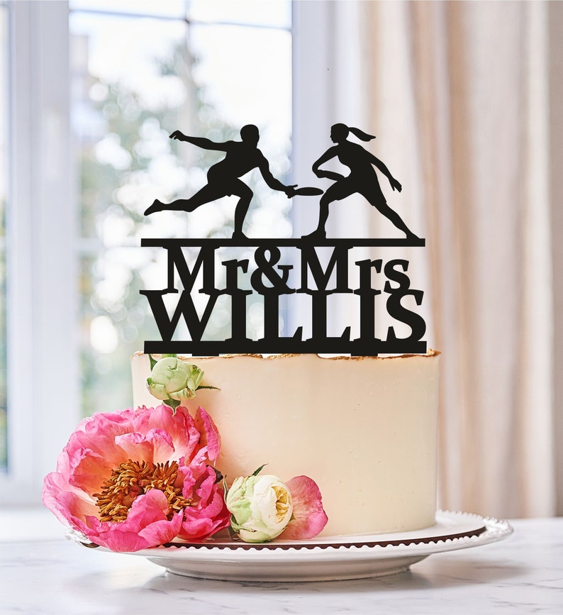 Peut inclure: Silhouette noire d'un couple jouant au frisbee avec le texte "Mr & Mrs Willis" en dessous. Le topper est plac&eacute; sur un g&acirc;teau blanc avec des fleurs roses et blanches.