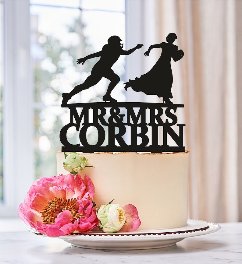 May include: Black silhouette cake topper of a bride and groom, the groom is in a football pose, the bride is holding a football. The topper says "Mr. & Mrs. Corbin" in black lettering.
