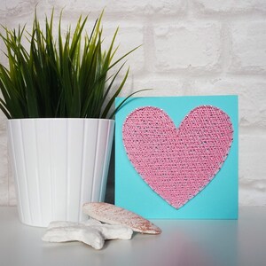 May include: A turquoise blue card with a pink heart made of string art. The heart is in the centre of the card. The card is sitting on a white surface.