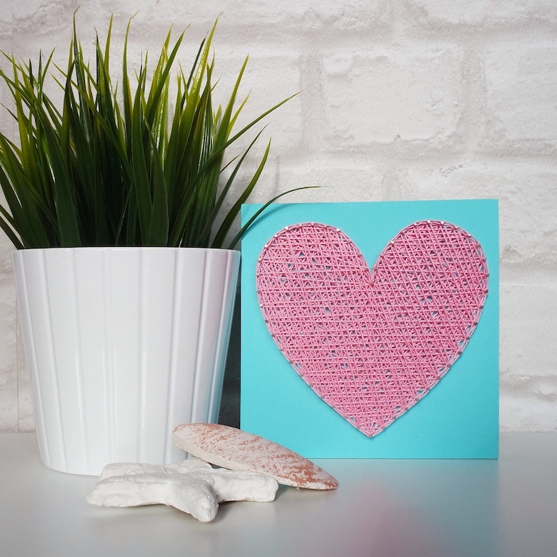 May include: A turquoise blue card with a pink heart made of string art. The heart is in the centre of the card. The card is sitting on a white surface.