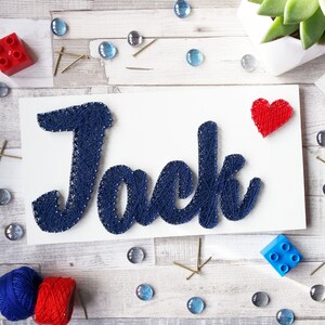 Puede incluir: Un letrero de madera blanco con el nombre "Jack" escrito en hilo azul. El hilo está envuelto alrededor de clavos para crear las letras. Un corazón rojo se encuentra en la esquina superior derecha del letrero.