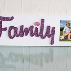 Könnte beinhalten: Ein weißes Holzschild mit dem Wort "Family" in lila glitzerndem Garn geschrieben. Ein kleines rosa Herz befindet sich über dem "i". Ein Foto von zwei Kindern ist in einem Rahmen auf der rechten Seite des Schildes.