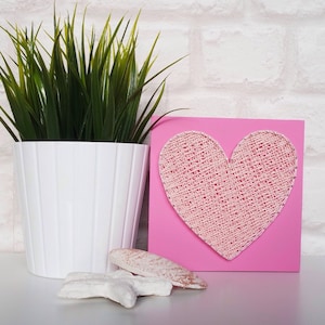 May include: A pink square wooden plaque with a white string art heart design. The heart is made of white string wrapped around nails hammered into the wood. The plaque is sitting on a white surface.