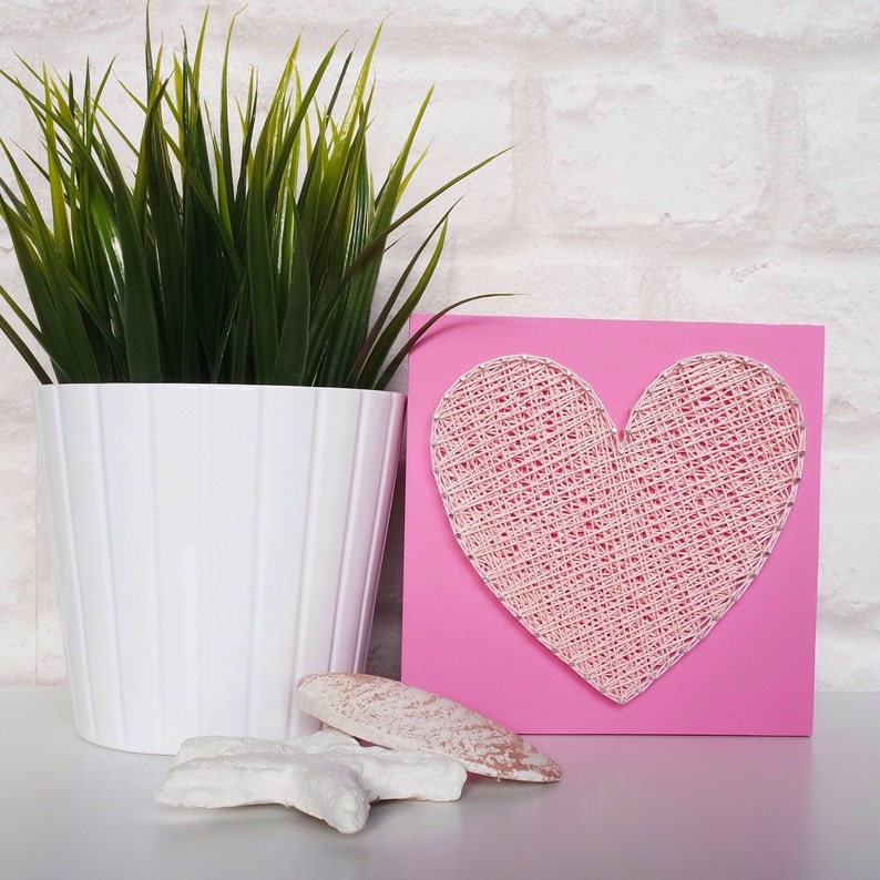 May include: A pink square wooden plaque with a white string art heart design. The heart is made of white string wrapped around nails hammered into the wood. The plaque is sitting on a white surface.