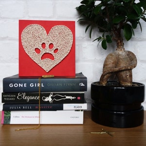 Könnte beinhalten: Eine rote quadratische Leinwand mit einem weißen String-Art-Herzdesign, das eine Pfote im Inneren zeigt. Die Leinwand liegt auf einem Stapel Bücher mit den Titeln "Gone Girl", "Elegance", "massive" und "The Woman in White".